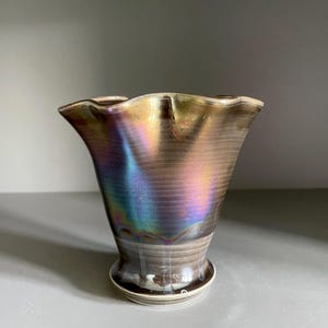 May include: An iridescent vase with a ruffled edge. The vase has a brown and gold metallic finish with hints of blue and purple. The vase sits on a small, round base. The vase is a decorative item.