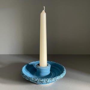 May include: A blue ceramic candle holder with a textured, crackled rim and a smooth central socket, holding a single, tall, slender, off-white taper candle. The base features a circular, shallow dish design with visible concentric rings.