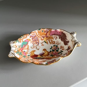 Puede incluir: Un plato ovalado de porcelana con borde festoneado y ribete dorado, que presenta un patrón colorido de faisanes, aves exóticas y motivos florales en tonos naranja, rosa, verde y marrón sobre un fondo blanco con detalles ornamentales.