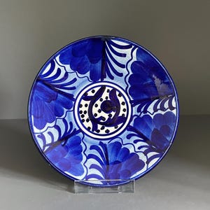 May include: A round ceramic plate featuring a hand-painted cobalt blue and white floral pattern. The design includes a central circular medallion surrounded by repeating petal motifs that extend towards the dark blue rim of the dish.