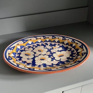 May include: Oval ceramic serving platter with a floral design. The platter features a white flower pattern with yellow centres on a blue background. The rim is orange, and the border has a yellow and blue geometric pattern.