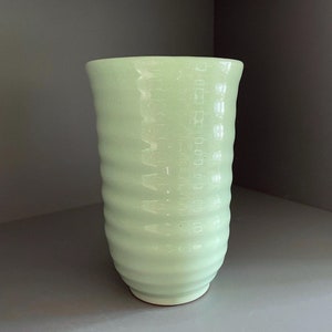 May include: A pale green ceramic tumbler with a textured, ridged design. The cup has a slightly flared rim and a rounded base. The surface has a subtle, glossy finish. The tumbler is empty and sits on a grey surface.