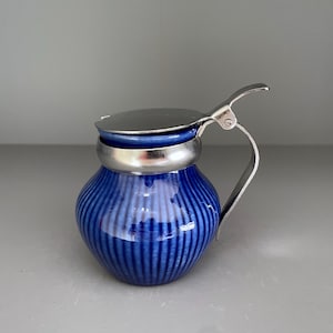 May include: A small, blue ceramic creamer with vertical ribbed detailing. It has a silver-colored metal rim, handle, and hinged lid. The creamer is a classic design, suitable for serving cream or milk.
