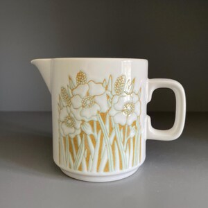 May include: A white ceramic creamer with a floral design in shades of gold and green. The creamer has a handle and a spout for easy pouring. The floral pattern is repeated around the body of the creamer.