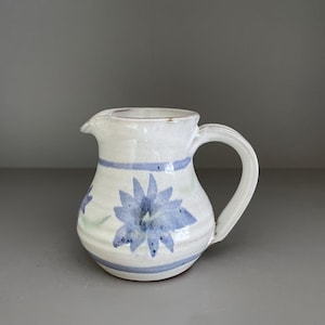Puede incluir: Una jarra de cerámica blanca con asa curva y boquilla. La jarra está decorada con diseños florales azules y rayas azules. El artículo está hecho a mano.