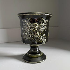 May include: A dark brown ceramic goblet featuring a raised relief design of a cherub playing a trumpet surrounded by grape clusters. The vessel has a wide rim, a tapered stem, and a circular base with a glossy finish and lighter green highlights.