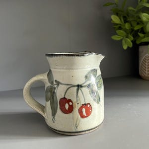 May include: A cream-coloured ceramic creamer pitcher featuring a hand-painted cherry motif with two bright red cherries and green leaves. The pitcher has a speckled texture, a curved handle, a small pouring spout, and a dark rim along the top edge.