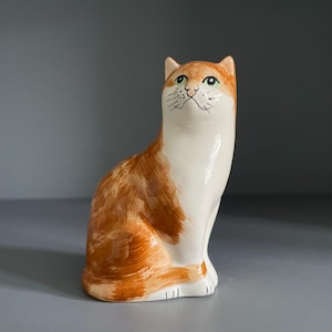 May include: A ceramic cat ornament in a seated position. The cat features orange and white markings, green eyes, and black whiskers. The figurine has a glossy finish and is placed on a grey surface.