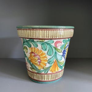 May include: A ceramic flower pot with a hand-painted floral design. The pot features a cream-colored base with vertical lines and a green rim. The floral pattern includes yellow, orange, pink, and blue flowers with green leaves.