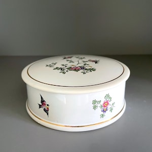 May include: A white, round ceramic trinket box with a lid. The box is decorated with floral and bird motifs in various colours, including pink, purple, and green. Gold trim accents the edges of the box.