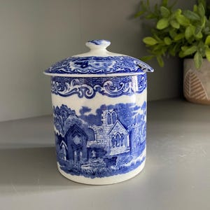 May include: A cylindrical ceramic jar with a lid, featuring a blue and white transferware pattern depicting architectural ruins and landscape scenes. The lid has a small knob handle and a notch for a spoon, with ornate scrollwork borders.