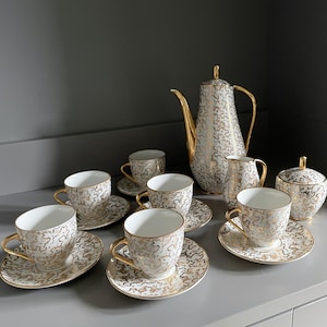Puede incluir: Un juego de té completo blanco y dorado con cafetera, crema, azucarero y seis tazas de té con platillos. El juego presenta un patrón de remolino dorado y blanco. Las tazas tienen asas y bordes dorados.