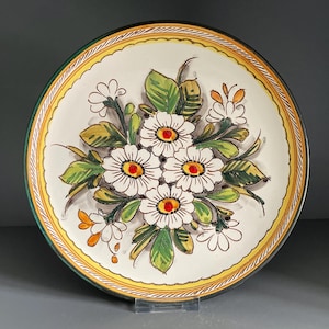 Puede incluir: Un plato de cerámica decorativo con un diseño floral pintado a mano. El plato tiene una base blanca con un borde amarillo y verde. El diseño central muestra margaritas blancas con centros amarillos y hojas verdes.