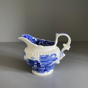 May include: A white ceramic cream jug featuring a blue transferware pattern depicting a landscape with trees and buildings. The jug has a scalloped rim, a curved handle with ornate detailing, and a rounded body with a fluted texture near the top.