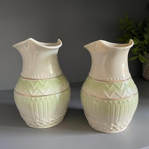 May include: Two ceramic jugs featuring a cream-coloured upper section and a pale green lower body. The jugs have a scalloped rim, a gold-toned band, and a textured surface with embossed chevron, vertical line, and floral patterns.