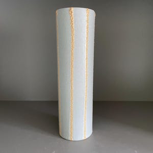 May include: A cylindrical vase with a light blue hue and vertical gold-coloured textured stripes. The vase is a decorative item, likely for holding flowers or as a standalone art piece. The vase is approximately 25 cm tall.