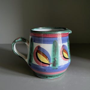 May include: A hand-painted ceramic jug with a handle and spout. The jug features a white base with colourful designs, including blue, red, and green stripes, and leaf-like shapes with yellow and brown accents. The jug is approximately 15 cm tall.