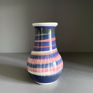 May include: A ceramic vase with a rounded body and a flared rim, featuring horizontal stripes in shades of deep blue, soft pink, and white. The surface has a smooth, glossy finish with a subtle marbled pattern throughout the colorful bands.