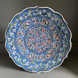 May include: A decorative ceramic plate with a scalloped edge. The plate features a vibrant blue background with intricate floral and geometric patterns in red, white, and green. The rim is white.