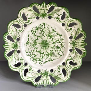 May include: A round ceramic plate featuring a white base with green floral patterns in the center. The plate has a decorative scalloped edge with cut-out details and green painted accents along the rim and the intricate border design.