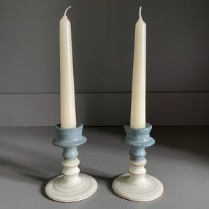 May include: Two ceramic candle holders with ivory candles. The candle holders are blue and white. The base is white, the middle section is a light beige, and the top is a light blue. The candles are tall and slender.