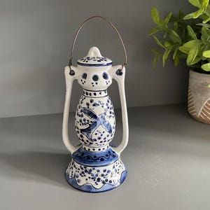 Könnte beinhalten: Eine weiße Keramiklaterne mit handgemalten blauen Blumen- und Vogelmotiven. Die Laterne hat einen abgerundeten Mittelkörper, einen ausgestellten Fuß und zwei seitliche Stützen, die oben durch einen dünnen, gebogenen Metalldrahtgriff verbunden sind.