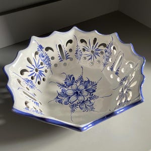 Puede incluir: Cuenco de cerámica blanca con borde festoneado y diseños florales azules. El cuenco tiene forma geométrica con ribete azul y detalles calados. El patrón floral está pintado a mano.