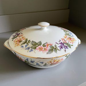 May include: A white ceramic serving dish with a lid, decorated with a floral pattern in pink, yellow, purple, and blue. The dish has gold trim and a small knob on the lid.