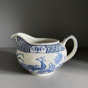 May include: Cream ceramic creamer with blue bird and floral pattern. The creamer has a spout and a handle. The design includes a blue border and a bird motif on the front. The creamer is a classic design.