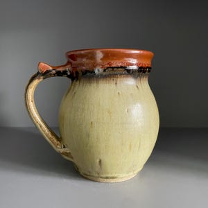 May include: A ceramic mug with a rounded body, featuring a glossy cream-coloured base that transitions into a dark brown drip pattern near the rim. The rim itself is a vibrant orange-red, and the mug includes a curved handle with a small thumb rest.