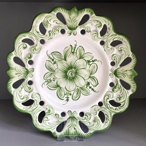 May include: A white ceramic plate featuring a hand-painted green floral design in the center. The plate has a scalloped, openwork border with intricate green scrollwork patterns along the edges, creating a decorative and detailed appearance.