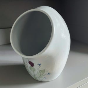 Pode incluir: Vaso de cerâmica branca com uma forma curva única. O vaso apresenta um design floral com flores roxas e azuis e folhas verdes. O vaso está sobre uma superfície cinza.