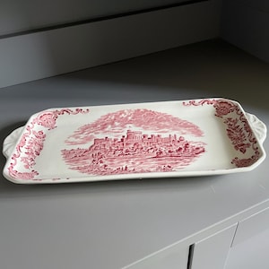 May include: A rectangular ceramic serving tray featuring a white base with a detailed crimson red transferware pattern. The center depicts a castle landscape, while the edges are decorated with ornate floral scrollwork and integrated side handles.