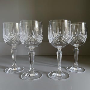 Puede incluir: Cuatro copas de vino de cristal transparente con patrones tallados en diamante. Las copas tienen un diseño clásico con base redonda, tallo largo y parte superior en forma de cuenco. Las copas están dispuestas sobre una superficie gris.