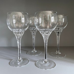 Puede incluir: Cuatro copas de vino de vidrio transparente con un diseño de tallo decorativo. Las copas están dispuestas en fila sobre una superficie gris.