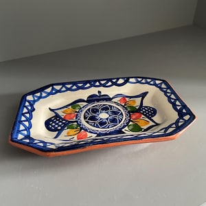May include: An octagonal ceramic tray with a cream-coloured base, featuring a hand-painted blue lattice border and a central floral motif in blue, yellow, green, and red. The underside of the tray reveals a natural terracotta clay finish.
