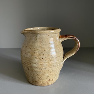 May include: A ceramic jug with a speckled beige and tan glaze, featuring a rounded body, a small pouring spout, and a curved handle with a dark brown accent. The surface has a textured appearance with subtle horizontal ridges around the base.