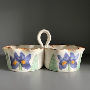 May include: A white ceramic serving dish with two compartments and a handle. Each section features a blue iris flower and green leaves. The rim has a brown, textured edge. Ideal for serving snacks or dips, it is a decorative piece.