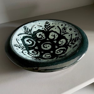 May include: A dark green ceramic bowl with a white glaze and a black floral design. The design features stylized flowers and swirls.