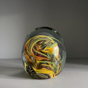 May include: A clear glass paperweight with swirling yellow, orange, and green internal designs. The paperweight has a rounded shape and a small label on the side. The glass is transparent, allowing the colorful patterns to be visible.
