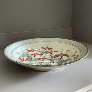 Puede incluir: Una fuente de cerámica con un diseño ancho y poco profundo. La superficie blanquecina está decorada con un estampado floral en rojo, azul y gris. El borde tiene una franja azul y un aspecto moteado y texturizado.