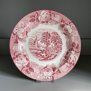 Puede incluir: Un plato de cerámica blanco con un patrón floral rosa y una escena central de un molino de viento y una granja en un entorno rural.