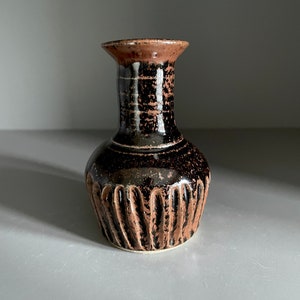 May include: A small, black and brown ceramic vase with a textured, ribbed base and a narrow neck. The vase has a white glaze with a speckled pattern.
