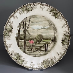 May include: A white ceramic dinner plate with a green and brown floral border. The center of the plate features a scenic image of a farmhouse in a rural setting with rolling hills and trees.