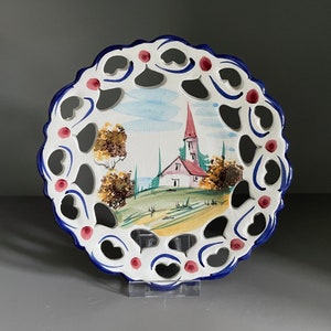 May include: A white ceramic plate with a blue and red border. The plate has a cut-out design and features a hand-painted scene of a church and trees.