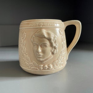 Puede incluir: Una taza de cerámica de color crema con una imagen en relieve de la cara de una mujer y el texto "JUNE 2nd 1953". La taza tiene un asa y un diseño floral.