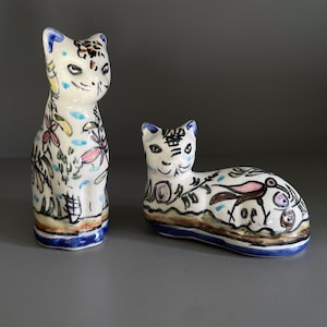 Puede incluir: Dos figuritas de cerámica de gatos con diseños florales y de pájaros pintados a mano. El gato más grande está de pie, mientras que el gato más pequeño está acostado. Ambos gatos son blancos con detalles azules, verdes, rosas y marrones.