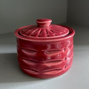 Könnte beinhalten: Ein kleines, rundes, rotes Keramikgefäß mit Deckel. Das Gefäß hat ein strukturiertes, geripptes Design am Körper und einen dekorativen Deckel mit einem kleinen Knopf. Das Gefäß wird wahrscheinlich zur Aufbewahrung kleiner Gegenstände verwendet.