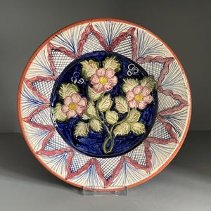 May include: A ceramic plate with a floral design. The plate features a central blue circle with pink flowers and green leaves. The rim has a red and white pattern. The plate is round and has a glossy finish.