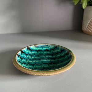 May include: An oval decorative dish with a teal and black swirled design. The dish has a textured, light brown rim. The dish is likely used for holding small items or as a decorative accent piece.